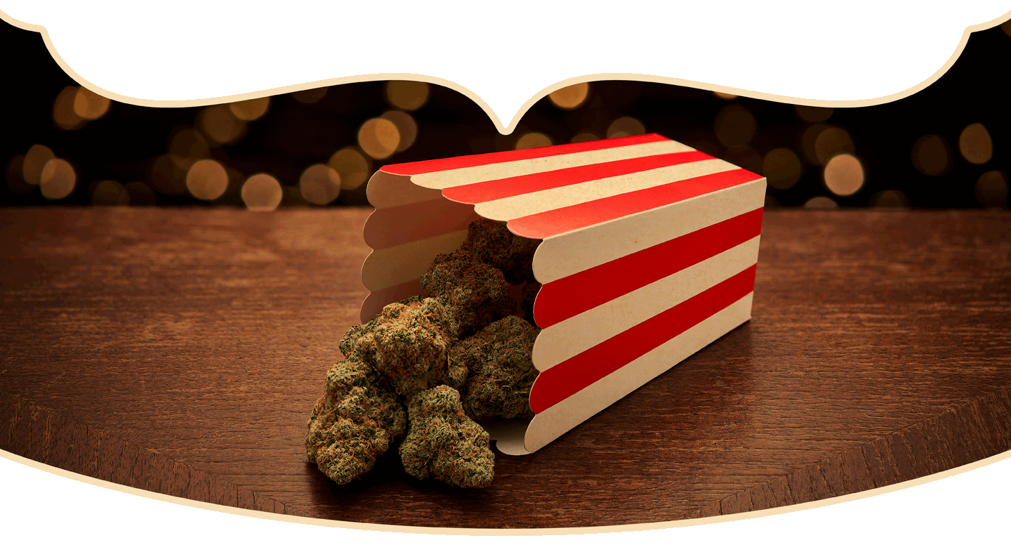 A red-and-white striped popcorn box tipped over on a wooden surface, with clusters of cannabis buds spilling out instead of popcorn. Blurred lights are visible in the background.